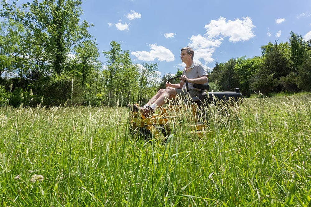 old man driving a zero turn mower on high grass
