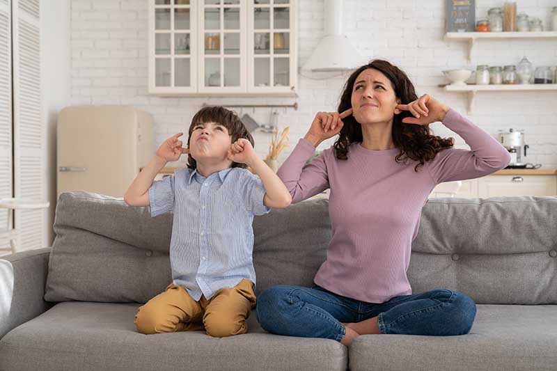 Frowning mom and son sit on couch with closed eyes and cover ears