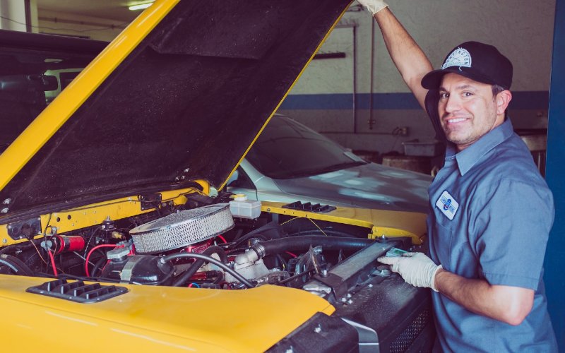 a car mechanic smiling in front of a yellow vehicle with an open hood