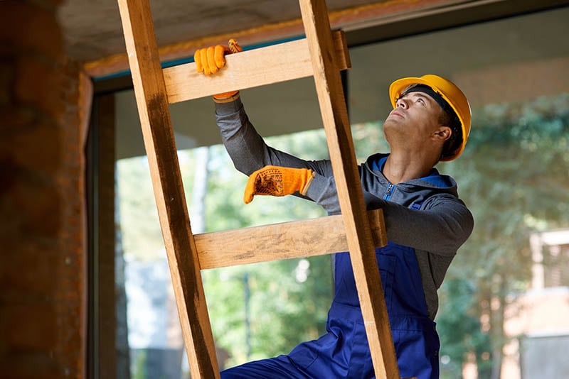Young male builder in overalls and hard hat lclimbing up the wooden ladder