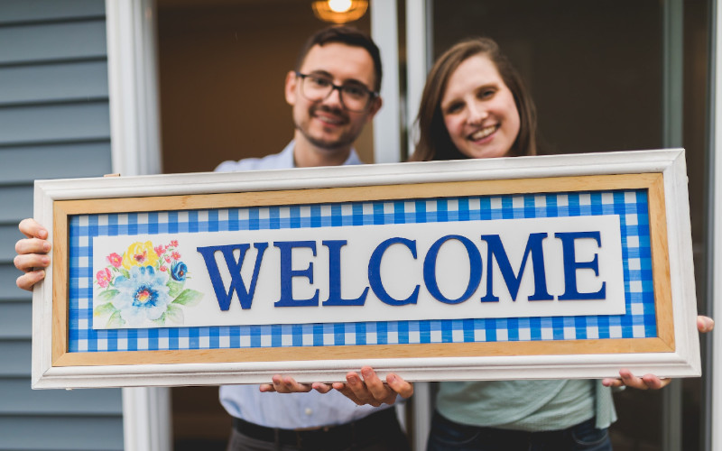 a couple holding up a framed welcome sign in front of house