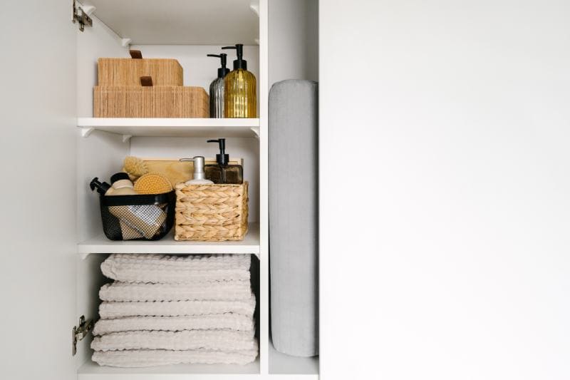 bath towels folded on shelf, wicker baskets with cosmetics bottles and massage brush in wooden cabinet