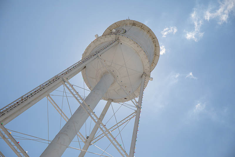 Photo looking up at a white water tower with a bright blue sky and back-lit by the sun