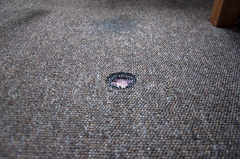 A burned hole in an old stained brown carpet