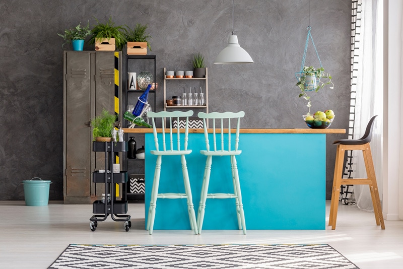 Wooden bar stools and blue dining table under lamp in room interior with grey wall
