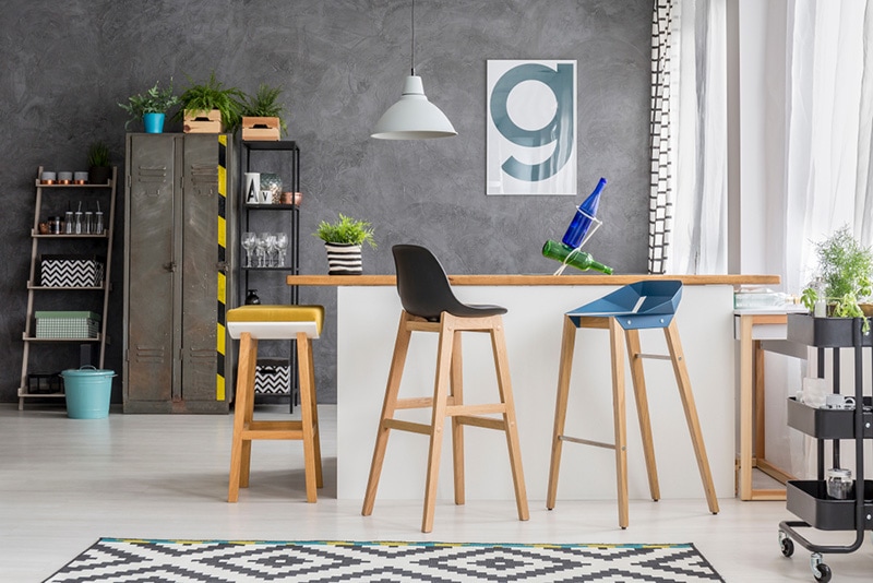 Wooden bar stools at kitchen island in bright kitchen with lamp and poster on concrete wall