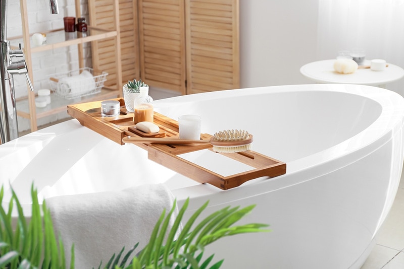 Wooden bathtub tray with different supplies and cosmetic products in light bathroom interior design