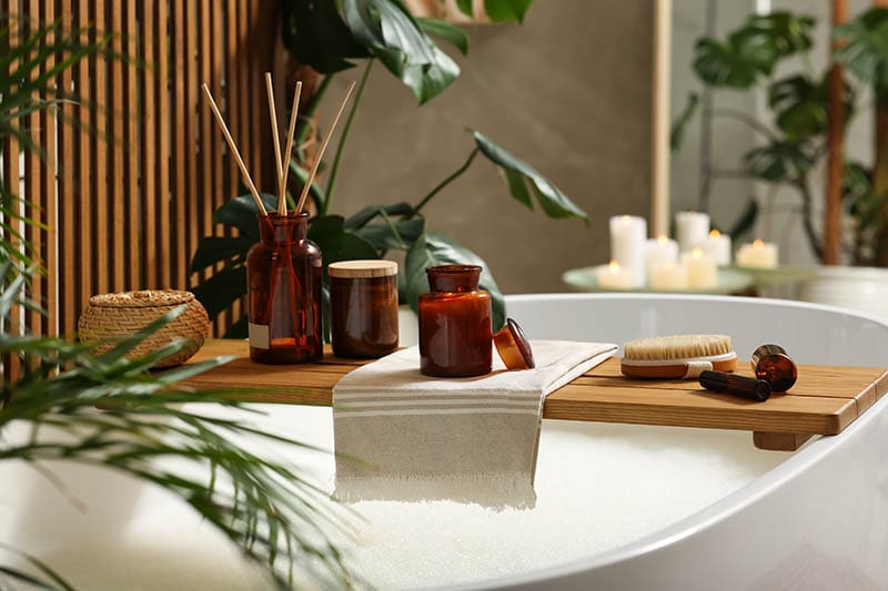Wooden bath tray with open book, candle and body care products on tub indoors